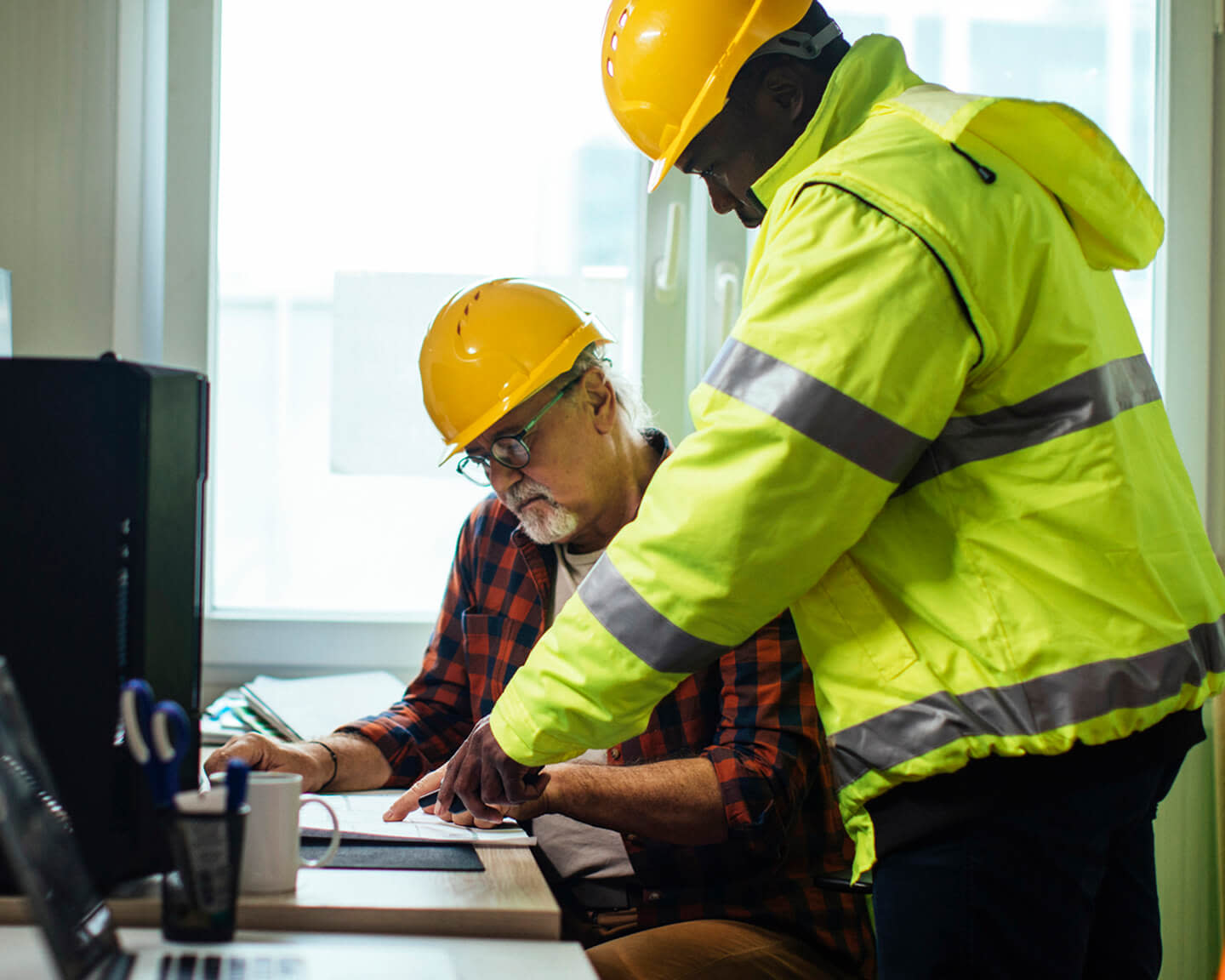 Two construction workers in helmets reviewing documents at a desk.