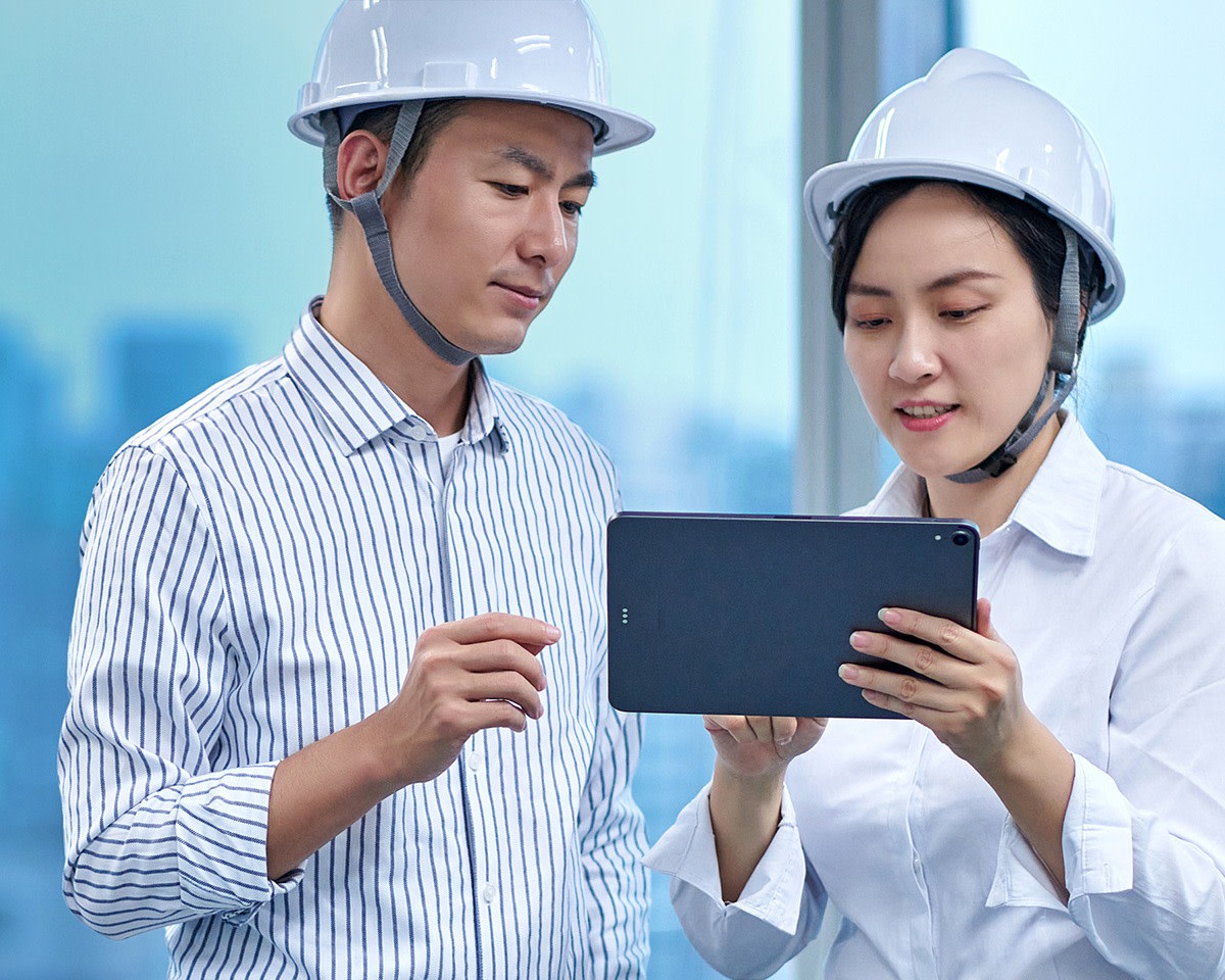 Two engineers in white helmets reviewing a tablet in an office.